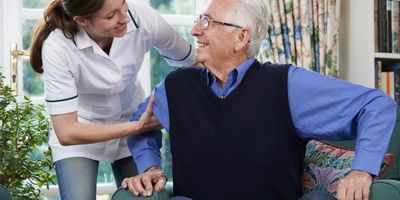 Kind caregiver assisting happy, dapper gentleman in blue to stand from armchair.