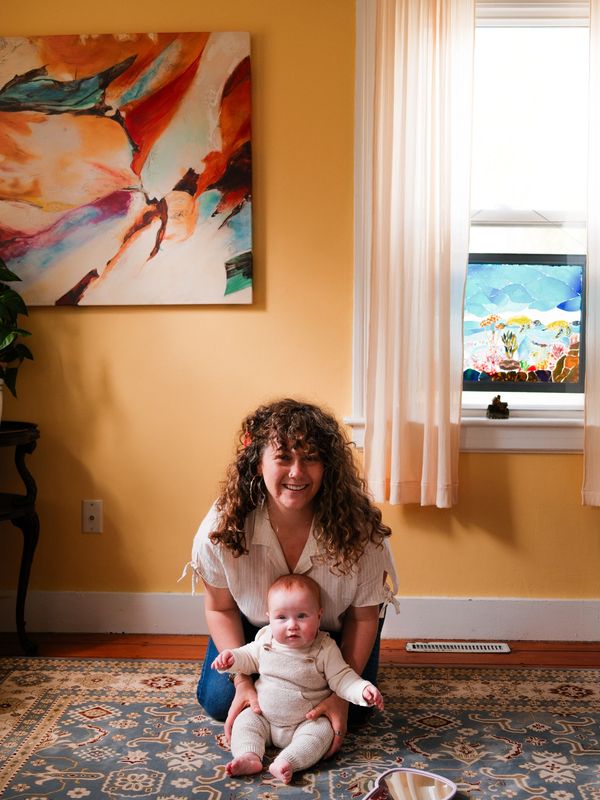 A smiling woman with curly hair holds a baby in a cozy, warmly lit room with artistic decor.