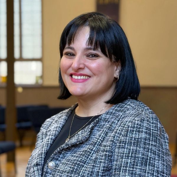 Smiling woman with black hair in a checkered blazer indoors.