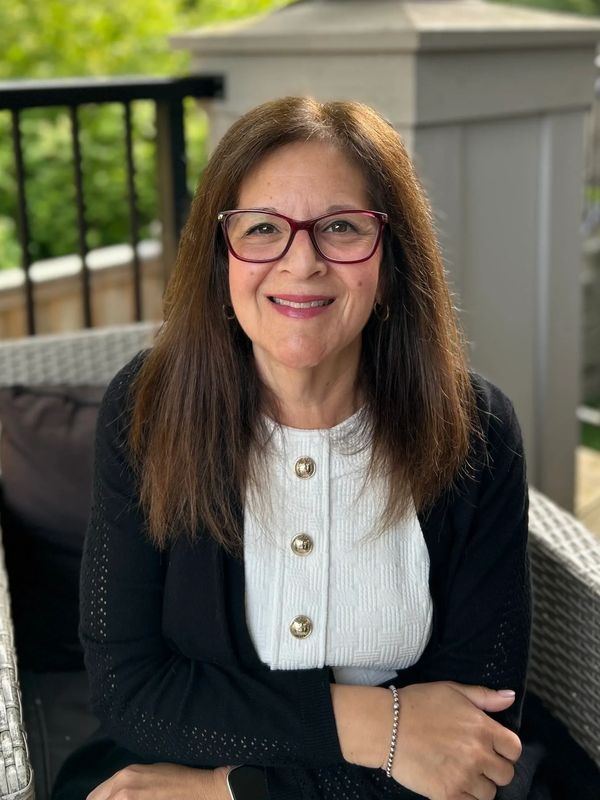Smiling woman with glasses sitting outdoors on a wicker chair.