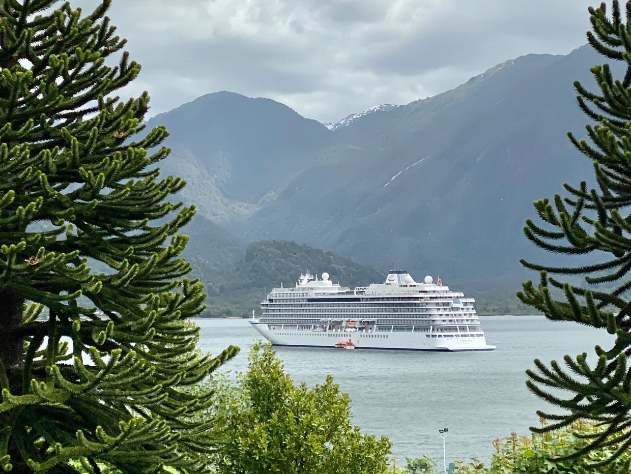 A cruise ship sails on a lake framed by pine trees and mountains under a cloudy sky.