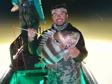 Smiling man holding a large striped fish on a boat at night.