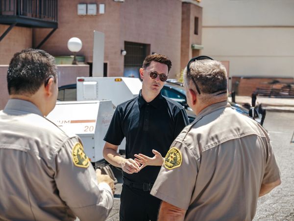 Man in black shirt talks to two uniformed officers outside.
