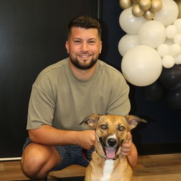A man smiling while holding a happy dog indoors with balloon decorations.