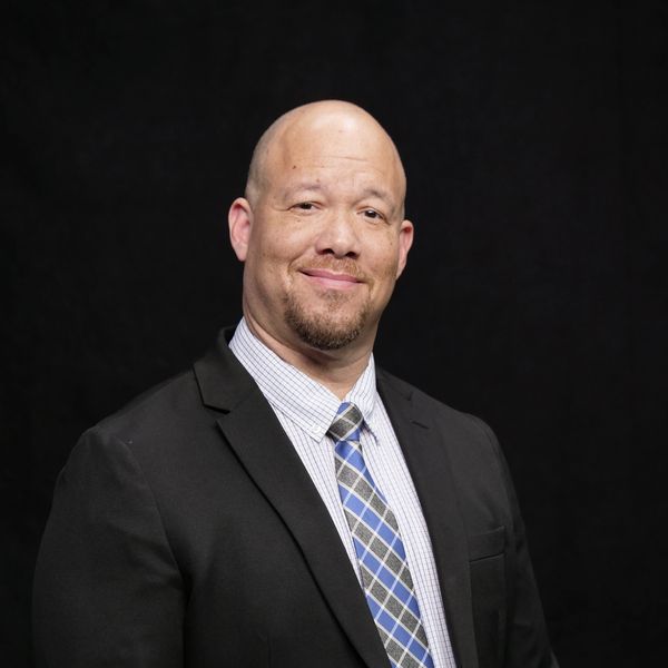 Smiling bald man in a suit and plaid tie against black background.