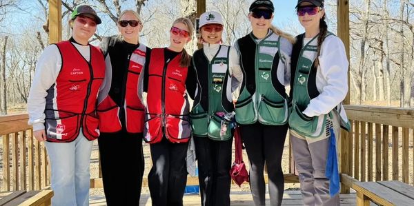 Six women in shooting vests posing outdoors on a wooden platform.