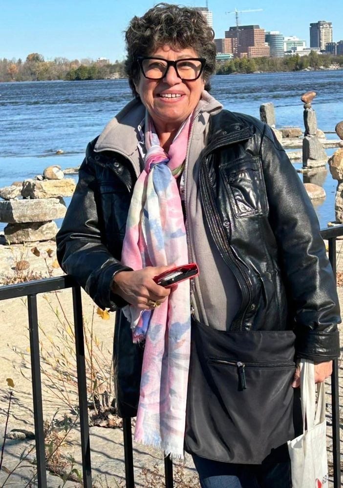 Smiling woman in a leather jacket and colorful scarf by a riverside with cityscape background.