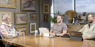 Three men engaged in a conversation at a wooden table in a cozy, art-filled room.