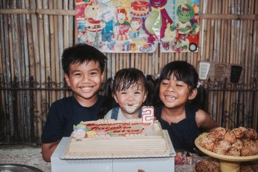 Three children smiling behind a birthday cake with a number 2 candle.