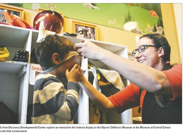 Female teacher helps a young child put on firefighter goggles at a children's museum.
