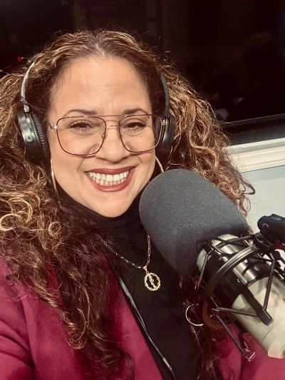 Smiling woman wearing headphones speaking into a microphone in a recording studio.