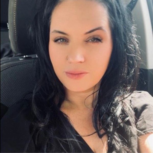 Woman with long black hair sitting in a car, looking at the camera.