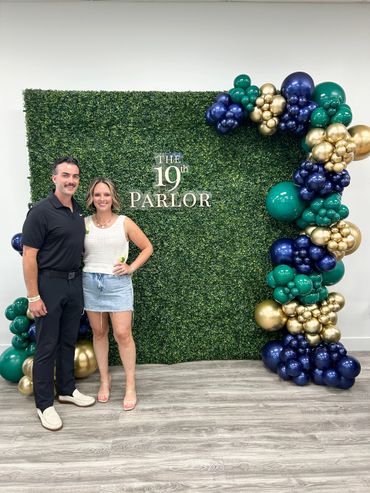 Couple posing in front of a green hedge backdrop with colorful balloon decorations.