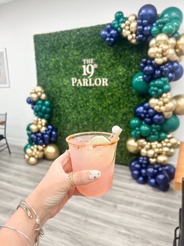 Hand holding a pink cocktail in front of a green wall with balloons and sign.