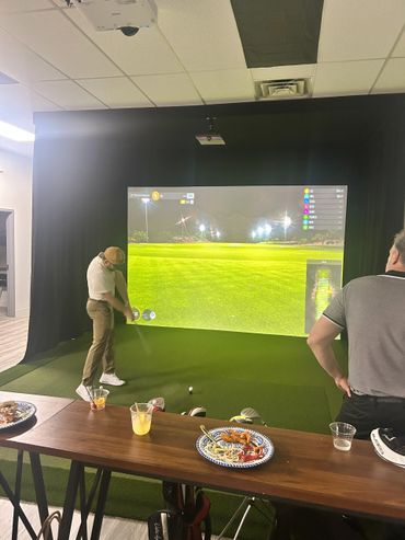 Two men playing indoor golf simulator with drinks and snacks nearby.