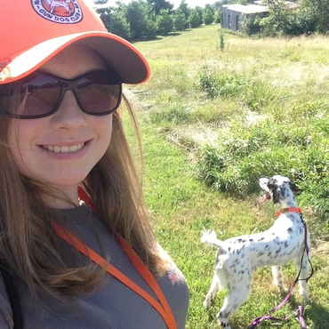 Smiling woman wearing sunglasses and a cap with her spotted dog outdoors.