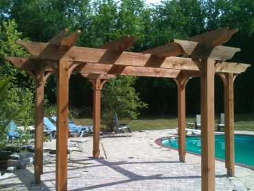 Wooden pergola by a poolside on a paved patio.