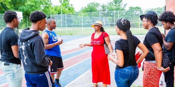 A woman in a red dress talks to a group of young people outdoors.