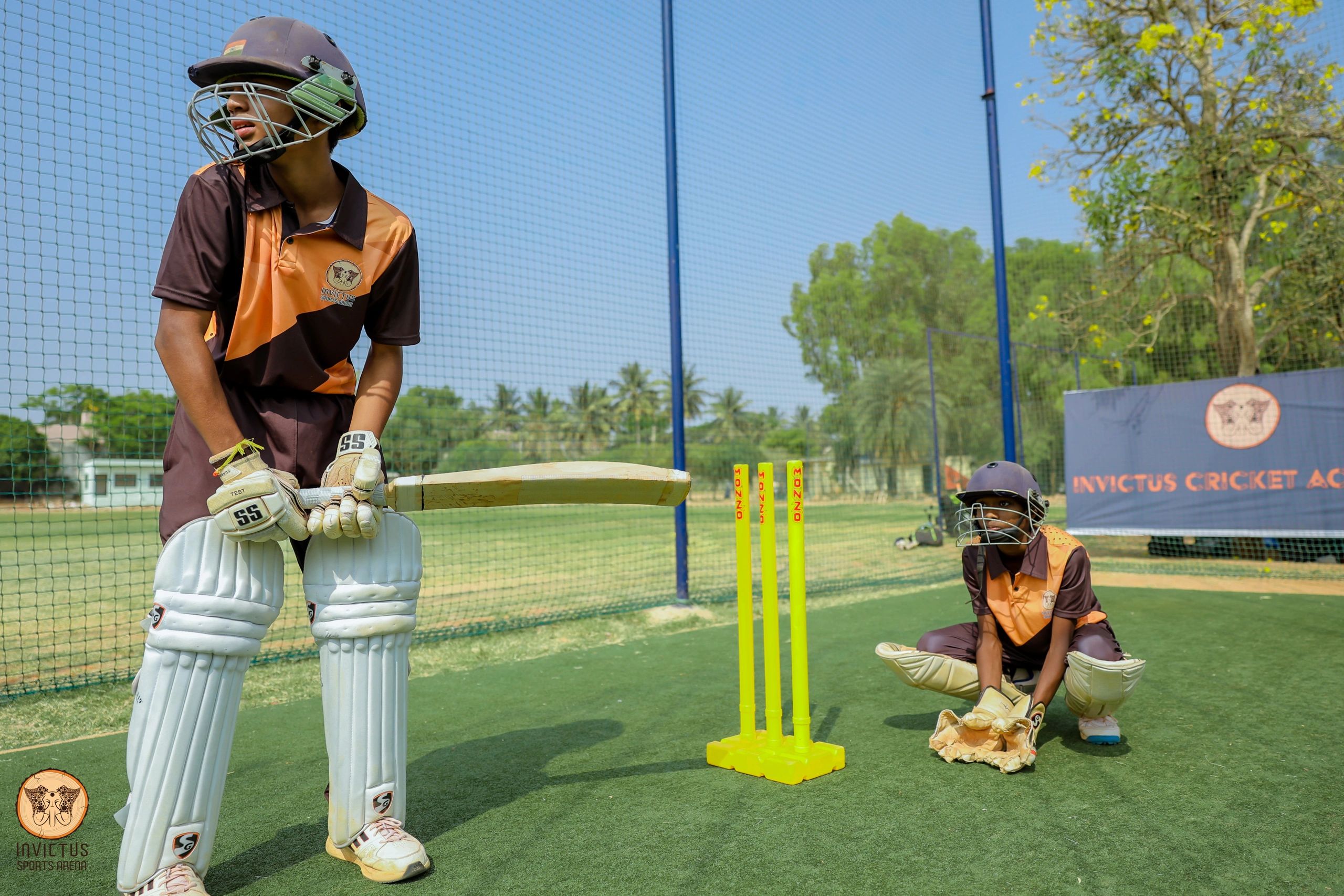 Two young cricketers in gear practicing on a green pitch under clear skies.