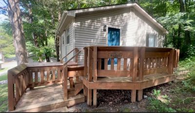Tan color cabin with bright blue door and large wooden deck surrounded by trees. 