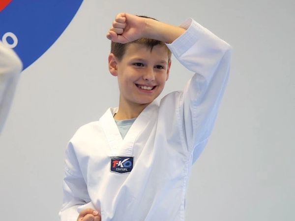 A boy in a white martial arts uniform smiling with his arm raised.