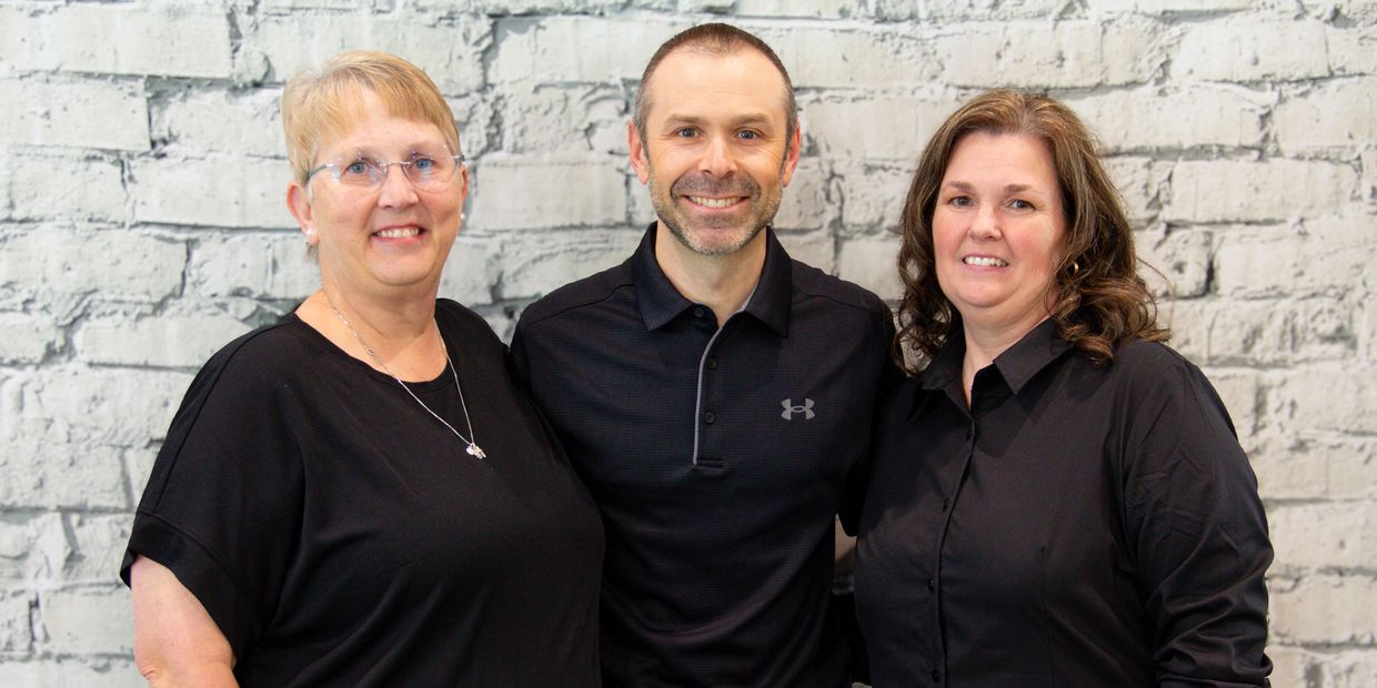 Three smiling adults in black shirts standing in front of a white brick wall.