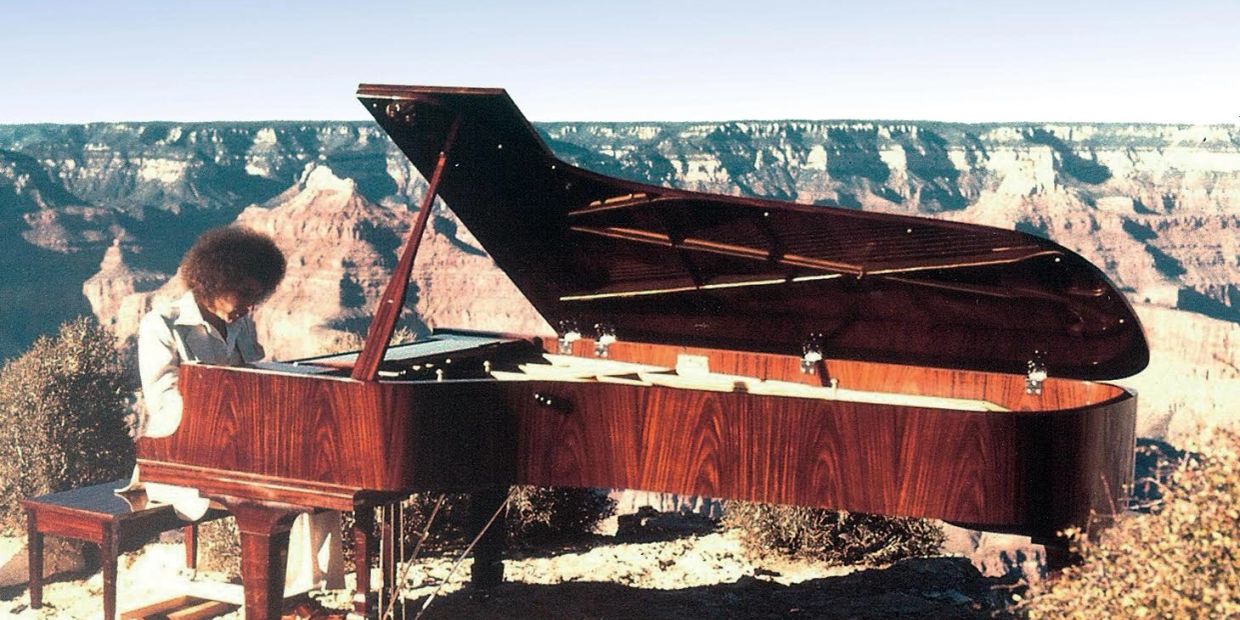 Person playing grand piano on a cliff with canyon backdrop.