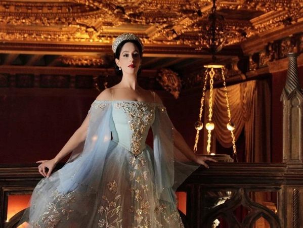 A woman in an elegant blue and gold gown stands in a grand, ornate room.