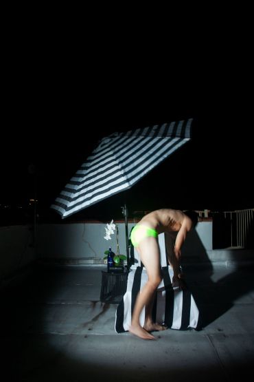 Person in neon green swimwear bending over under a striped umbrella at night.
