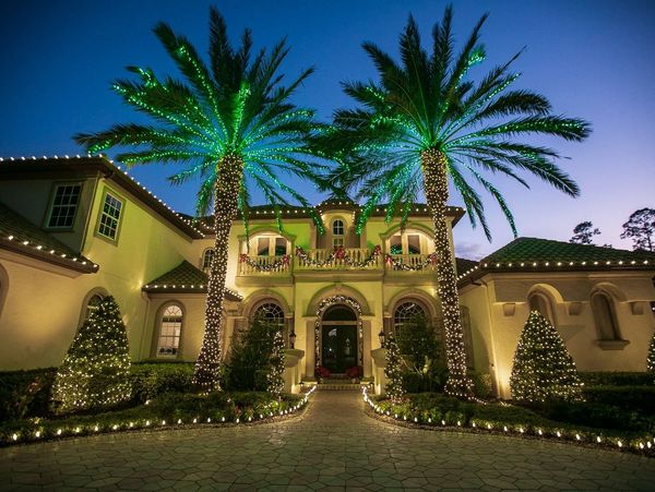 Luxury home decorated with festive lights on palm trees and bushes at dusk.