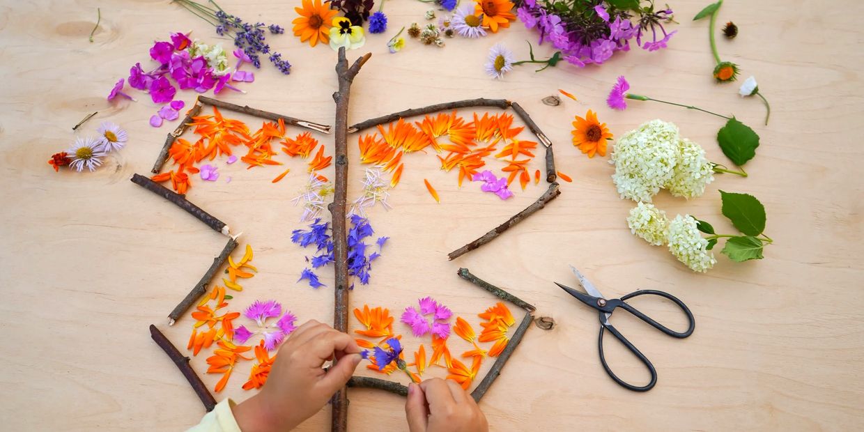 Bird's-eye view of a child's hands arranging fresh flowers and sticks into a butterfly shape. 