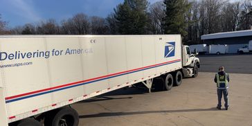 A USPS delivery truck parked near a building with a worker standing nearby.