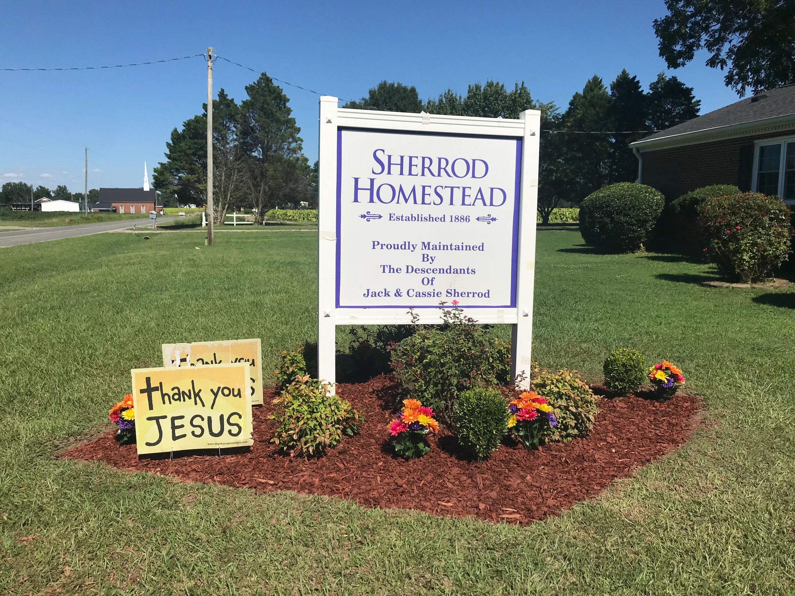 Sign for Sherrod Homestead with colorful flowers and a 'thank you JESUS' sign nearby.