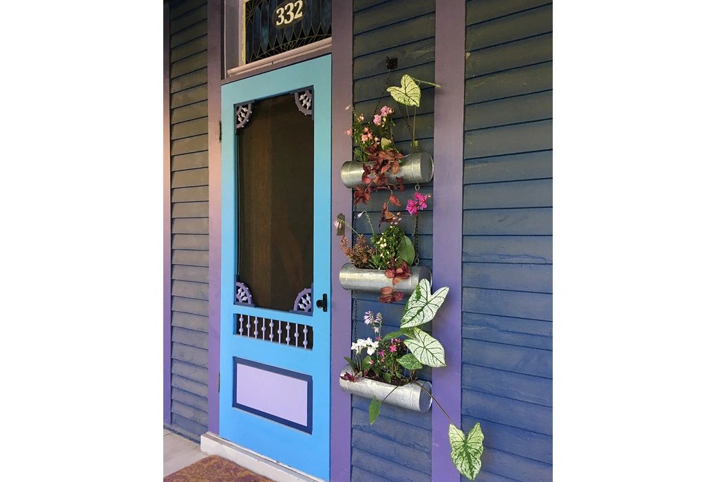 Front porch of Victorian home painted shades of purple and blue. Decorative plant holder near door.