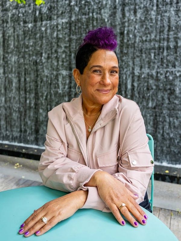 Smiling woman with purple hair and nails sitting at a table.