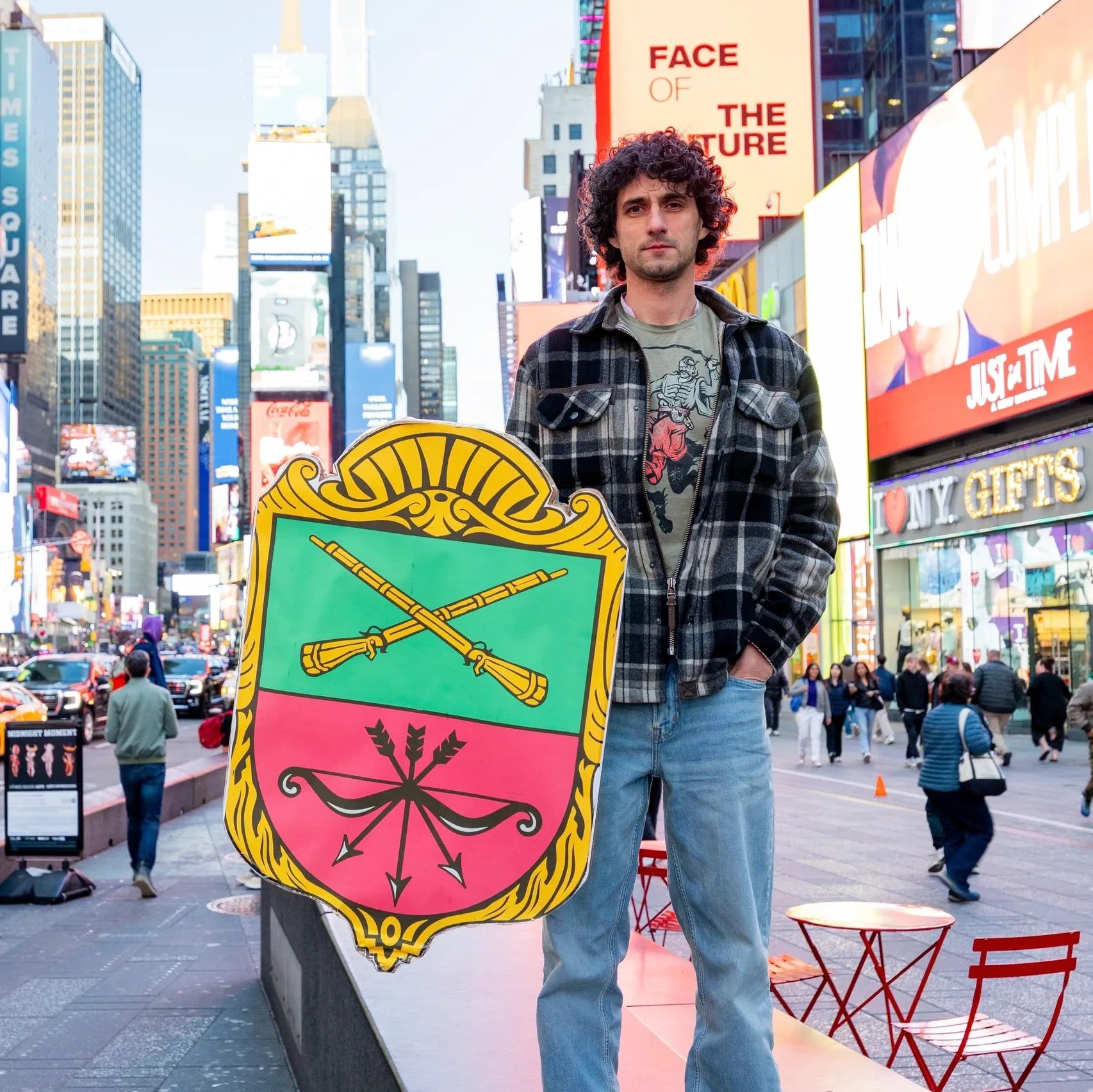 Vsevolod Myrnyi standing in Times Square holding a colorful shield with crossed rifles and arrows.