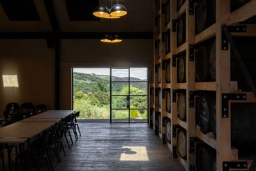 Interior of a rustic room with wooden tables, chairs, and a large window showing green hills.