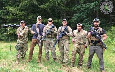 Group of six armed men in military gear posing outdoors in a forested area.