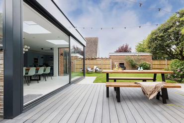 Modern backyard with sliding glass doors, wooden deck, picnic table, and green lawn under a cloudy sky.