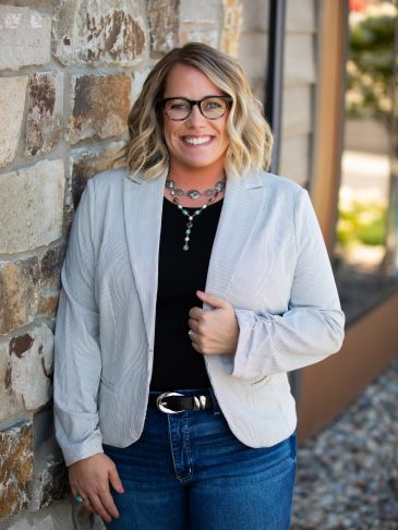 Smiling woman wearing glasses, a gray blazer, and jeans standing by a stone wall.