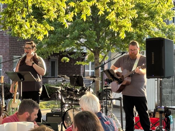 Live outdoor music performance with a singer and guitarist under green trees.