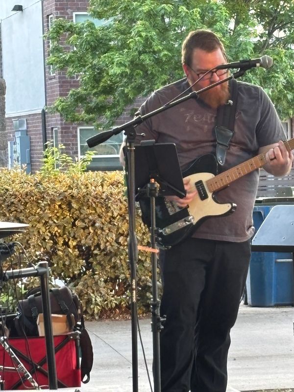 Man playing electric guitar outdoors with microphone stand in front.