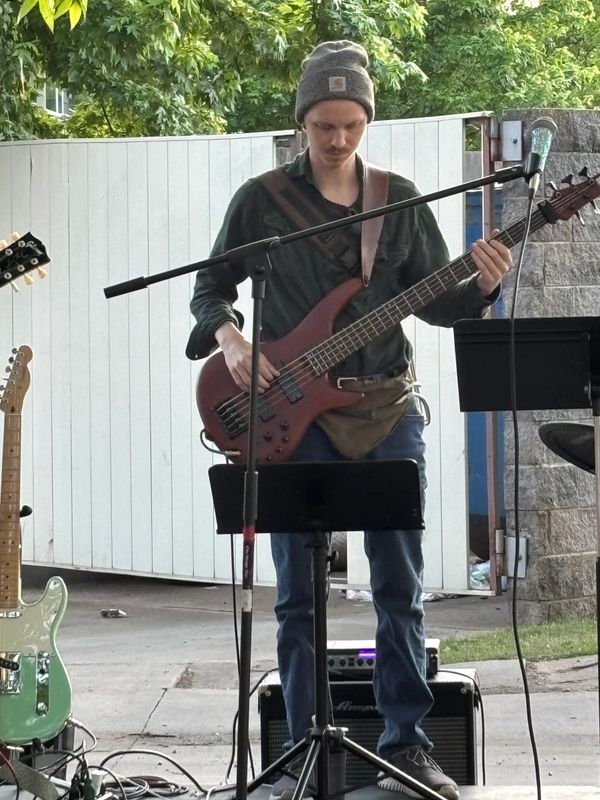 Musician playing bass guitar at an outdoor event with a microphone stand in front.