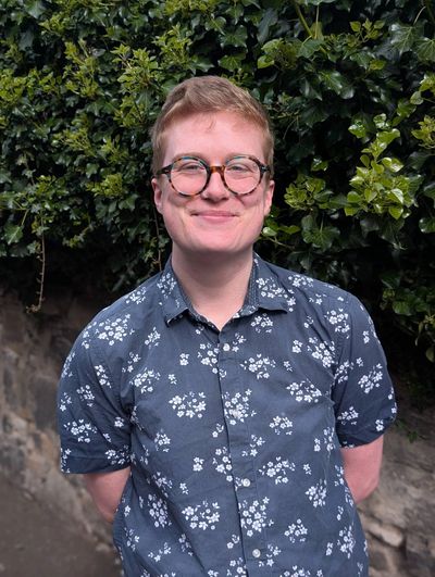 A smiling white person with blond hair and glasses, wearing a blue top. They stand in front of leave