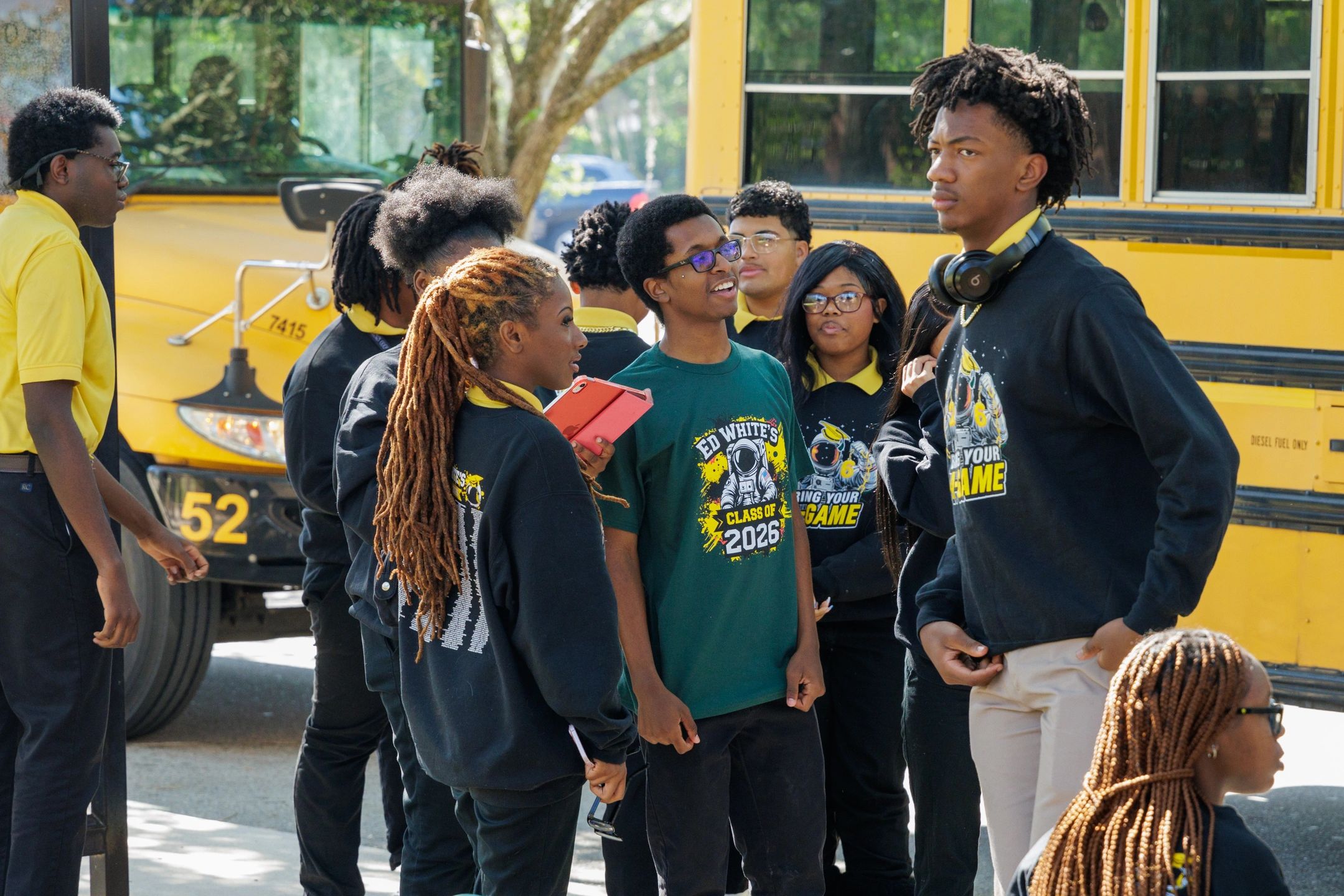 High school students chatting near a yellow school bus, some wearing Ed White's Class of 2026 shirts.