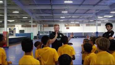 Coach instructing children in yellow shirts at an indoor sports facility.