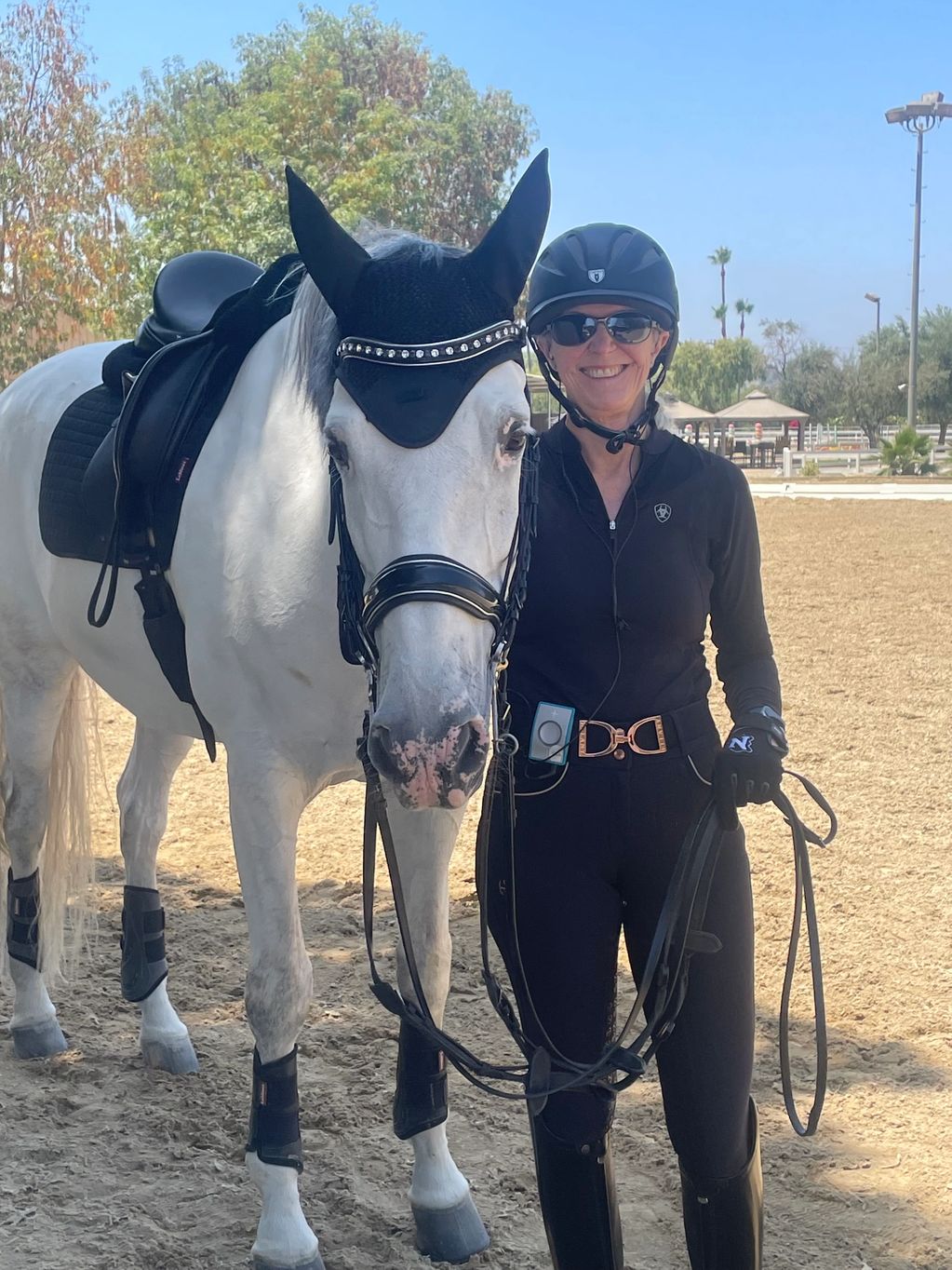 A smiling woman in equestrian gear stands next to a white horse with black tack.