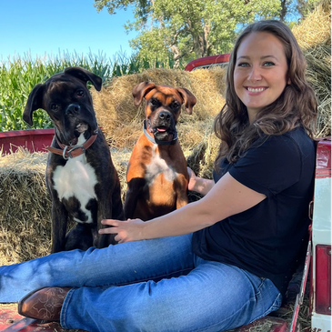 A woman sitting on hay with two boxer dogs, smiling outdoors.