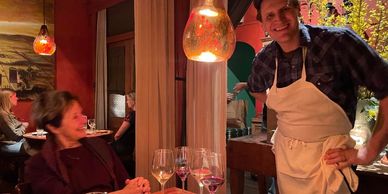 A smiling waiter poses with a seated woman in a warmly lit restaurant.