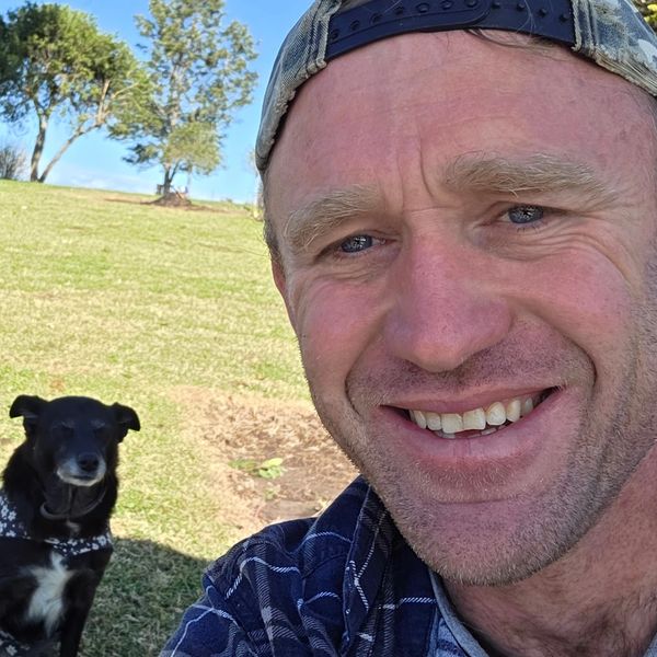 A man smiling outdoors with a black dog in the background.